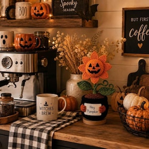 Könnte beinhalten: Eine gehäkelte orangefarbene Kürbisblume mit grünen Blättern und Stiel, eingetopft in einen schwarzen Behälter mit einem weißen Schild, auf dem "Hey Pumpkin" steht. Der Artikel steht auf einer Holzoberfläche neben einer Kaffeemaschine.