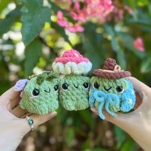 May include: Three small, handmade crochet plushies featuring soft green yarn textures. The left figure has purple wings, the center is a mushroom with a pink cap, and the right figure wears a brown hat and blue bow. Each has black eyes and a smile.