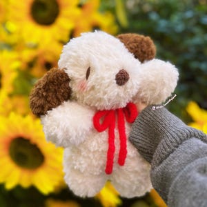 May include: A plush toy shaped like a puppy with white fur, brown ears, and a small brown nose. The puppy has pink blush on its cheeks and a bright red yarn bow tied around its neck. The text @alice.inwoollyland is visible on the side.