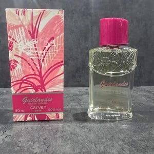 Op de afbeelding: Een heldere glazen parfumfles met een roze dop en een bijpassende doos. De doos heeft een roze en wit bloemenpatroon met de woorden "Guirlandes" en "carven paris". De fles bevat 50 ml parfum.