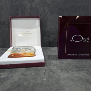 Op de afbeelding: Een parfumfles in een witte doos met een bordeauxrode rand. De fles is helder met een amberkleurige vloeistof. De doos is open, naast een bordeauxrode doos met de tekst "J'ai Osé" en "Parfums Guy Laroche Paris".