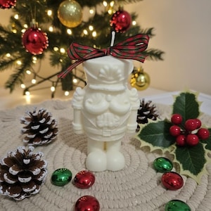 Può includere: Una candela color crema a forma di soldato schiaccianoci, ornata da un fiocco a righe rosse e verdi. La candela è circondata da decorazioni natalizie, pigne e agrifoglio, con un albero di Natale decorato sullo sfondo.