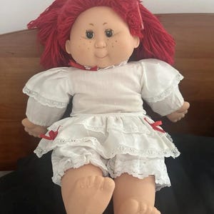 May include: A vintage doll with red yarn hair, freckles, and a white dress with lace trim. The doll is wearing a white top and ruffled shorts, with red ribbon accents. The doll's arms and legs are made of a soft material.
