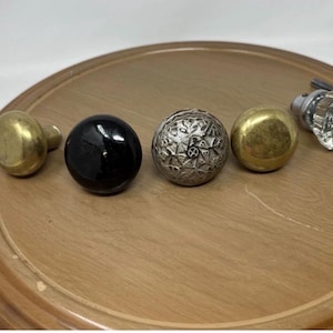 May include: An assortment of decorative cabinet knobs in various styles. Includes a brass knob, a glossy black knob, a silver floral design knob, a gold knob, and a clear faceted glass knob. All knobs are displayed on a wooden surface.