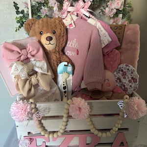May include: A white wooden crate filled with baby items, including a teddy bear, pink sweater, and headbands. The crate is decorated with pink flowers, butterflies, and the name "Ezra" in pink letters. A giraffe teether is also visible.