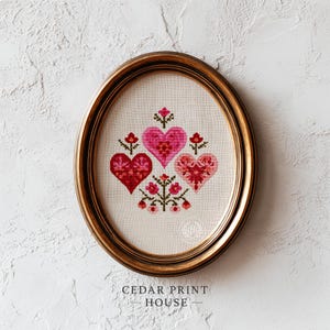May include: An oval framed cross-stitch featuring three hearts and floral accents in shades of red and pink. The artwork is set against a neutral background and framed in a gold-toned oval frame. The text "CEDAR PRINT HOUSE" is at the bottom.