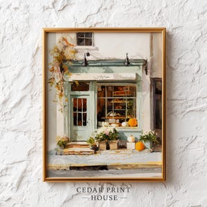 May include: A framed art print depicting a storefront with a light green door and window frame. The scene features orange pumpkins, white flowers, and autumn foliage. The building has a white facade and a small sign above the door. The artwork is enclosed in a gold-colored frame.