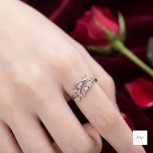May include: A silver-toned open ring featuring a delicate, swirling branch-like design. One end of the band is adorned with a small, round red gemstone and a clear stone, while the other end features a cluster of three clear, sparkling stones.