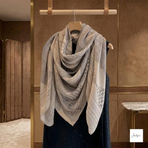 May include: A large, square scarf in a light beige hue featuring a dark gray geometric line pattern. The scarf is draped over a wooden hanger and displays various grid, stripe, and angular designs across its surface. The fabric appears soft and lightweight, hanging in loose folds that showcase the intricate, repeating linear motifs throughout the entire piece.