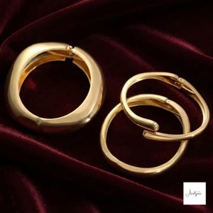 Puede incluir: Tres pulseras de aro articuladas de metal en tono dorado con acabado pulido. Una pulsera es más gruesa y ancha que las otras dos, que son más finas y delgadas. Cada pieza cuenta con un mecanismo de bisagra seguro para abrir y cerrar.