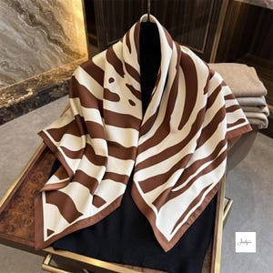 Puede incluir: Un pañuelo cuadrado con un patrón abstracto similar al de una cebra en marrón oscuro y crema. El pañuelo está drapeado sobre una prenda negra, mostrando su textura suave y bordes limpios. El diseño consiste en formas orgánicas fluidas.