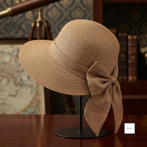 May include: A tan straw hat with a rounded crown and a wide brim. The hat features a matching tan fabric band wrapped around the base of the crown, which is accented with a large, decorative bow tied on the side with two long, flowing fabric tails.