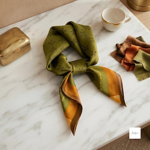 Peut inclure: Un foulard carré vert olive orné d'un motif géométrique répétitif, avec une large bordure dans des tons jaune moutarde et marron. Le foulard est noué, présentant une finition satinée lisse et des bords soigneusement finis.
