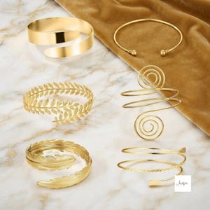 May include: A collection of six gold-toned metal bracelets featuring various designs including a smooth cuff, a thin wire bangle with ball ends, a leaf-patterned band, a spiral wire cuff, a feather-shaped wrap, and a textured spiral wire bracelet.