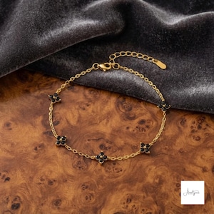 May include: A gold-tone chain bracelet featuring four evenly spaced flower-shaped charms, each set with four small black stones. The bracelet includes a delicate link chain, a lobster claw clasp, and an adjustable extension chain with a small metal tag.