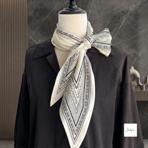 May include: A cream-colored square scarf featuring a black paisley and geometric border pattern, tied in a knot around the neck of a mannequin wearing a black long-sleeved shirt. The scarf displays a repeating teardrop motif and fine lines.