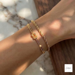 May include: Two gold-toned bracelets worn together. One bracelet features a delicate rope chain design, while the other is a smooth, flat chain that forms a central knot detail. Both pieces have a polished, reflective finish that catches the light.