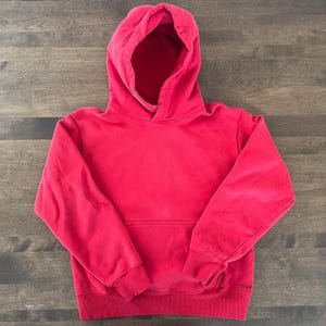 Può includere: Una felpa con cappuccio rossa con tasca a marsupio. La felpa è realizzata in un materiale morbido e ha un cappuccio con coulisse. Le maniche hanno polsini a coste. La felpa è di colore uniforme e si trova su una superficie di legno.