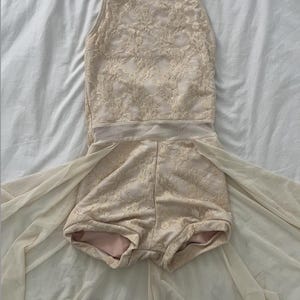 Puede incluir: Maillot de baile beige con cuello halter y shorts. El maillot está cubierto con un delicado patrón de encaje. La tela transparente se extiende desde los shorts, creando un efecto de falda fluida. La prenda se muestra sobre una superficie blanca.