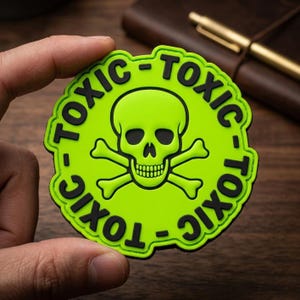 May include: A round, neon green patch with a black border and the word "TOXIC" repeated around the edge. The center features a lime green skull and crossbones graphic. The patch is held by a hand.