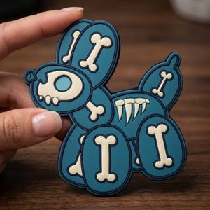 May include: A teal and white patch in the shape of a balloon dog with bone details. The patch has a dark blue outline and is held by a hand. The dog's head has a skull-like design.