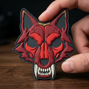 May include: A red and black patch featuring a stylized wolf skull design. The patch has a detailed, three-dimensional appearance with sharp teeth and gold accents on the ears. The overall design is bold and striking.