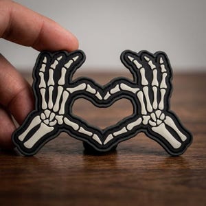 May include: A black and white skeleton hand heart-shaped patch. The patch features a black border with white bone details forming a heart shape. The patch is held up by a hand.