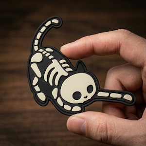 May include: A black cat-shaped patch with a skeleton design in off-white. The patch features a playful cat pose with a curved tail and outstretched paw. The design is a fun, spooky accessory.