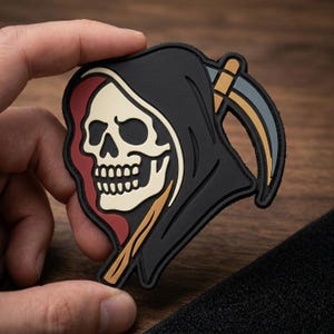 May include: A black patch featuring a Grim Reaper skull with a black hood, a beige skull, and a scythe with gray and gold accents. The patch has a red border and is held against a wooden background.