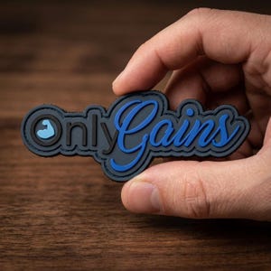 May include: A black and blue patch with the words "Only Gains" in a stylized font. The patch features a small blue arm icon and is held in a hand against a wooden background.