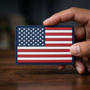 May include: A rectangular patch featuring the American flag. The flag design includes white stars on a blue background in the upper left corner and red and white stripes. The patch has a dark blue border.