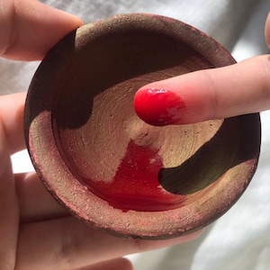 Puede incluir: Un pequeño cuenco redondo de color terracota con un borde dorado, lleno de líquido rojo brillante. Un dedo se sumerge en el líquido rojo, mostrando el color y la textura. El cuenco se sostiene en una mano.