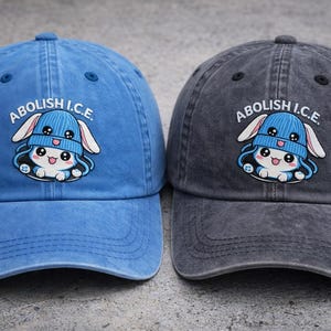 Può includere: Due cappellini da baseball, uno blu e uno grigio, ciascuno con la scritta "ABOLISH I.C.E." sopra un coniglietto dei cartoni animati con un cappello blu. I cappellini hanno una visiera curva e un cinturino regolabile.