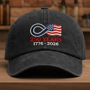 May include: A black baseball cap with an embroidered design. The design features an infinity symbol, an American flag, and the text "250 YEARS 1776-2026" in red and white. The cap is sitting on a wooden surface.