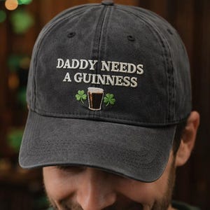 May include: Dark gray baseball cap with the embroidered text "DADDY NEEDS A GUINNESS" above a graphic of a pint of Guinness and two shamrocks. The cap is made of a soft, worn fabric.