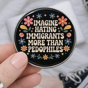 Op de afbeelding: Een ronde, zwarte button met de tekst "IMAGINE HATING IMMIGRANTS MORE THAN PEDOPHILES" in het wit, omringd door kleurrijke bloemen. De button wordt vastgehouden door een hand, met een vage achtergrond met een schoen.