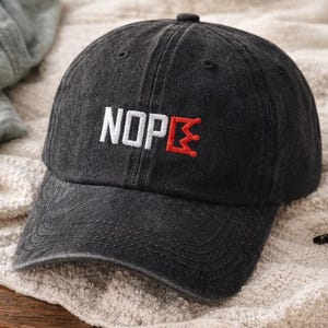 Pode incluir: Boné de beisebol preto com a palavra "NOPE" bordada em branco e um desenho de coroa vermelha. O boné tem uma aba curva e um fecho de tira de tecido. O chapéu é feito de um material macio e desgastado.