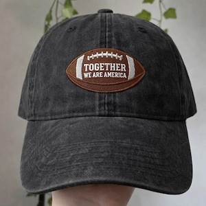 May include: A dark grey baseball cap with a worn look. It has a brown football-shaped patch with the white text "TOGETHER WE ARE AMERICA". A casual, everyday accessory.