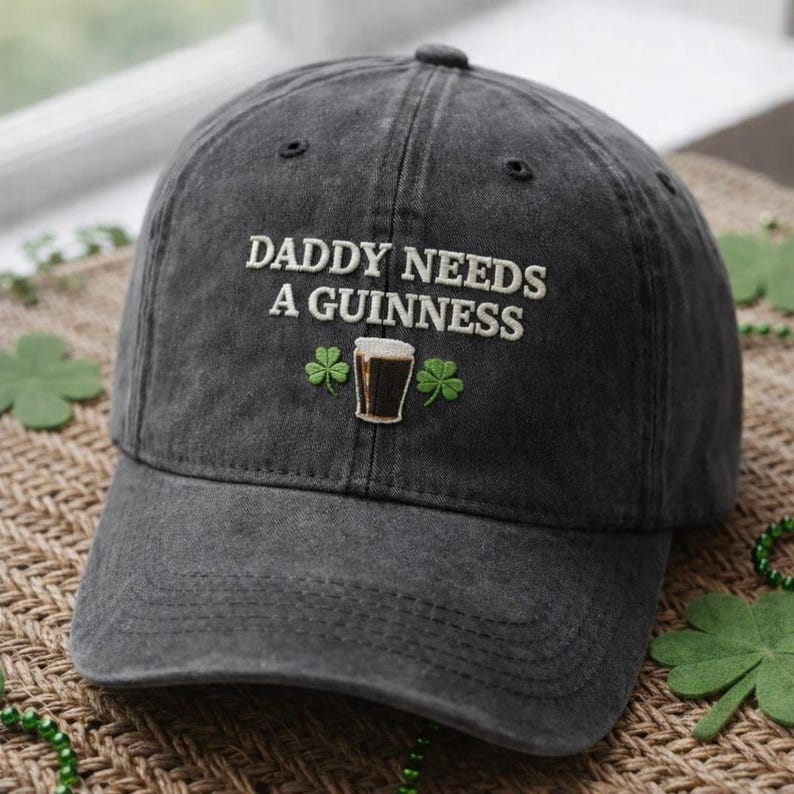May include: A charcoal gray baseball cap with the embroidered text "DADDY NEEDS A GUINNESS". The design includes a small embroidered image of a pint of Guinness and two shamrocks. The cap is a classic six-panel design.