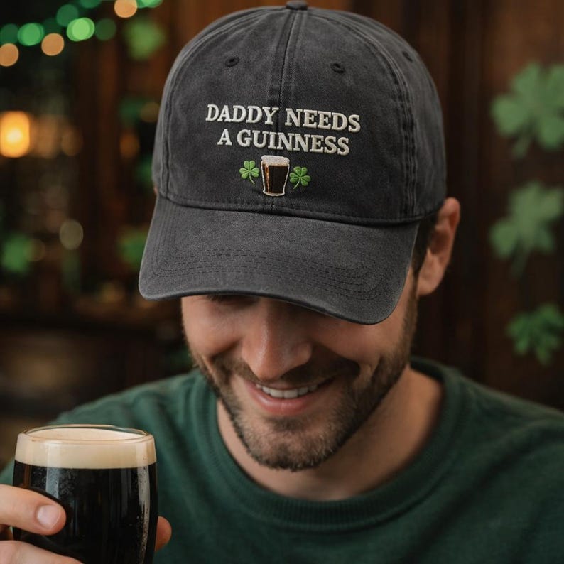 May include: Dark gray baseball cap with the embroidered text "DADDY NEEDS A GUINNESS" above a graphic of a pint of Guinness and two shamrocks. The cap is made of a soft, worn fabric.