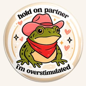 May include: A round button featuring a cartoon frog wearing a pink cowboy hat and a red bandana. The text "hold on partner, I'm overstimulated" is written around the frog. The design includes hearts and stars.