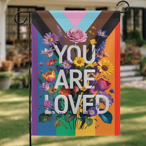 Op de afbeelding: Een tuinvaandel met een kleurrijk bloemendesign en de woorden "YOU ARE LOVED" in het wit. De vlag heeft een regenboogachtergrond met strepen en een zwarte metalen ophangstang.