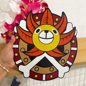 Puede incluir: Decoración de pared circular de madera con un diseño de sol de dibujos animados. El sol tiene una cara amarilla, rayos naranjas y un diseño de hueso blanco en la parte inferior. El fondo es marrón, rojo y blanco. La obra de arte se sostiene en una mano.