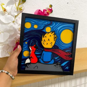 Puede incluir: Una obra de arte enmarcada y en capas que representa una escena de El Principito. La obra presenta al Principito con un zorro rojo, sobre un cielo estrellado con una gran luna amarilla. El marco es negro.