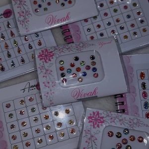 May include: Multiple rectangular packages of adhesive bindi stickers featuring various circular and teardrop shapes in assorted colors. Each white card displays the brand name Vivah or Ananya with floral patterns and contains rows of decorative face gems.