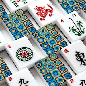 May include: A close-up shot of a Mahjong set with white tiles featuring various symbols and characters in red, green, and black. The tiles are arranged on a blue and gold patterned surface.