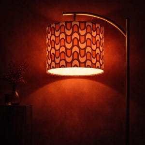 May include: A mid-century modern style floor lamp with a curved brass arm. The lampshade is cylindrical, featuring a repeating pattern of orange and dark brown curved lines on a cream background. The lamp casts a warm, inviting glow.