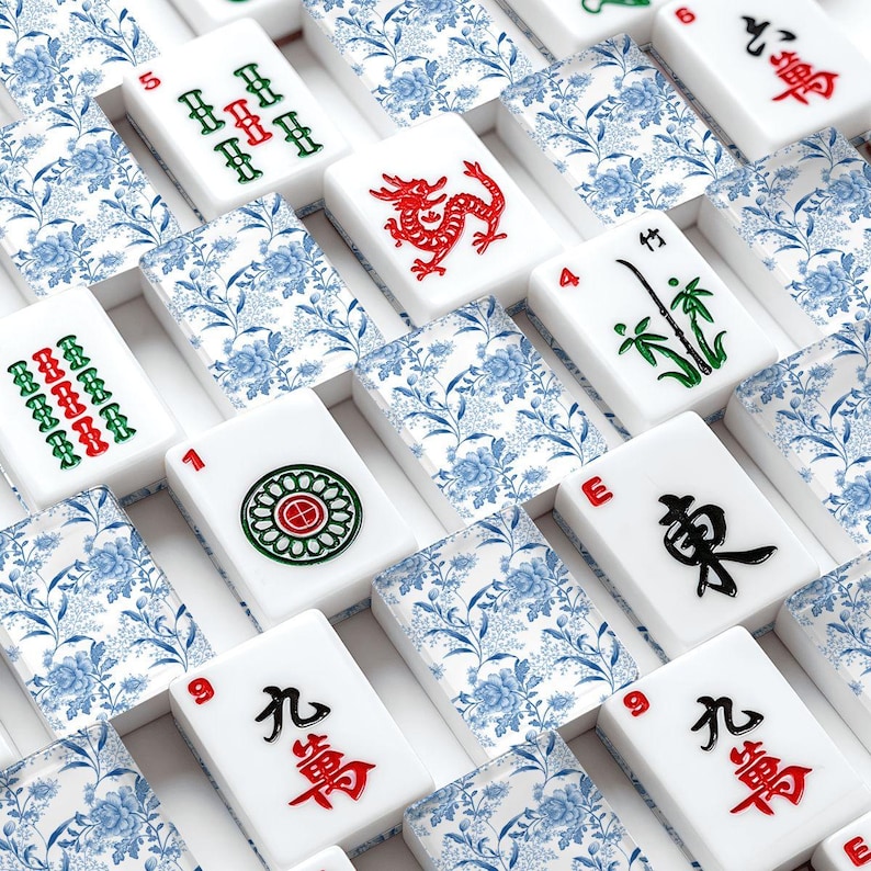 May include: A close-up shot of a Mahjong set. The white tiles feature red, green, and black symbols and numbers. The tile holders have a blue floral pattern. The set includes a red dragon tile.