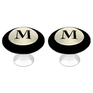 May include: Two round cabinet knobs featuring a black outer ring and a central brushed silver circular inlay. Each knob is engraved with a prominent black capital letter M in a serif font. The knobs have a silver-toned base and stem.