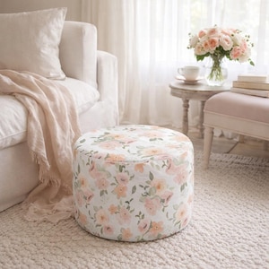 May include: A round ottoman with a floral pattern in shades of pink and peach on a white background. The ottoman is placed on a white rug, and the scene includes a white sofa, a side table with flowers, and a small pink bench.